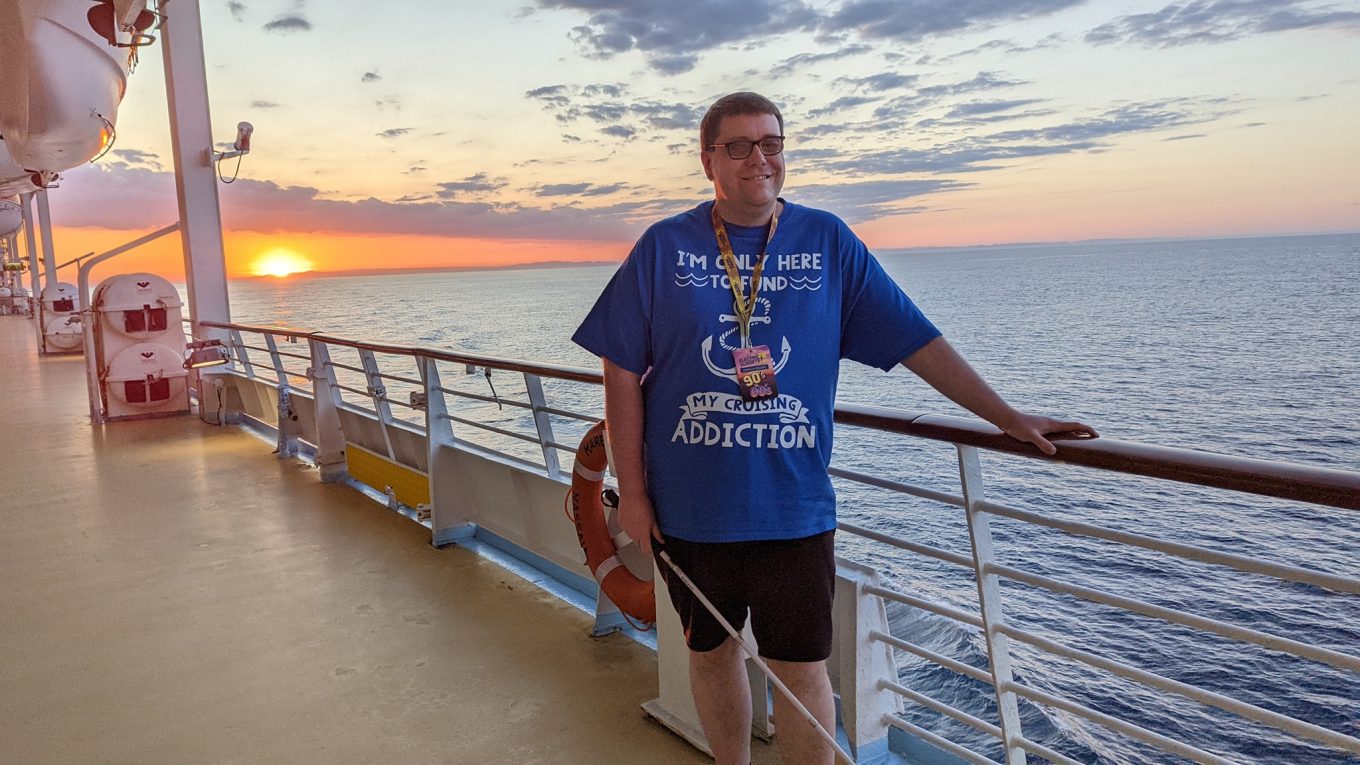 Chris, wearing a cruise themed t-shirt, stands in front of the sunset on the promenade deck of Marella Discovery 2.