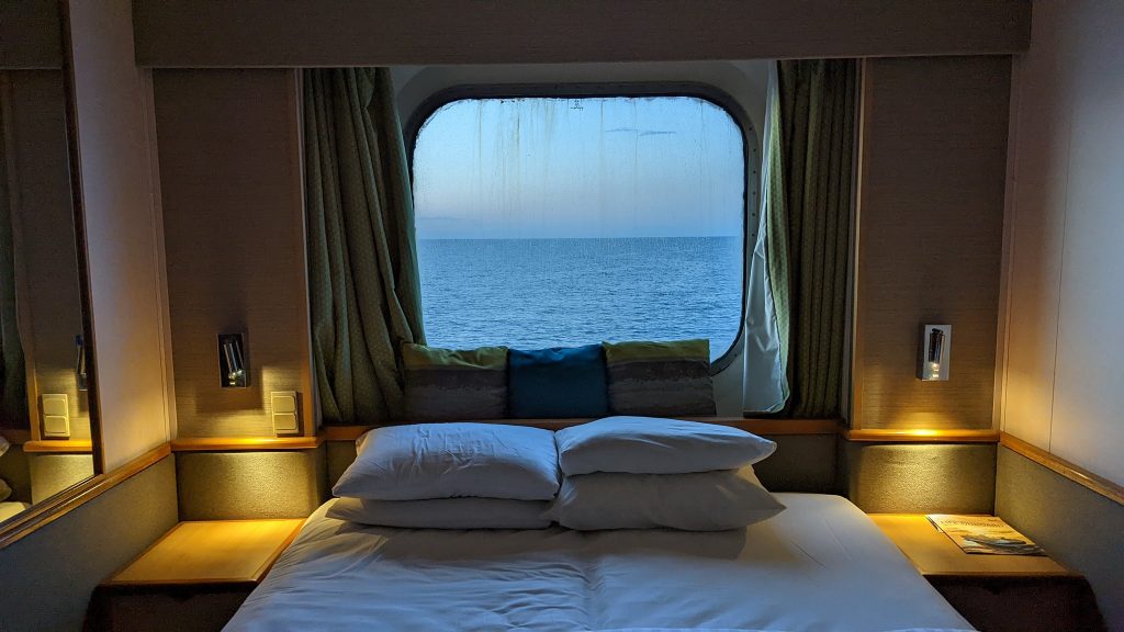 A view of the bed in my cruise ship cabin, with the sea visible through the window as the sun goes down.
